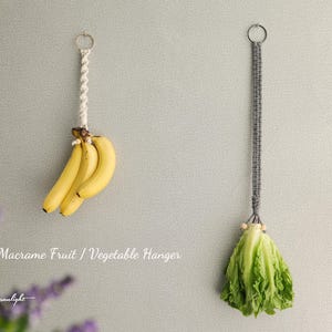 May include: Two macrame fruit and vegetable hangers. One is white with bananas hanging from it. The other is grey with a head of lettuce hanging from it. The text "Macrame Fruit / Vegetable Hanger" is written below the hangers.