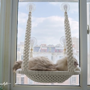 Może przedstawiać: Biała hamak dla kota z makramu jest zawieszona na oknie za pomocą przyssawek. Kot leży w hamaku i patrzy przez okno.