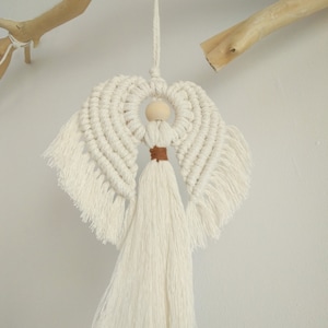 May include: White macrame angel ornament with a wooden bead head and brown tassel. The angel is hanging from a white cord.