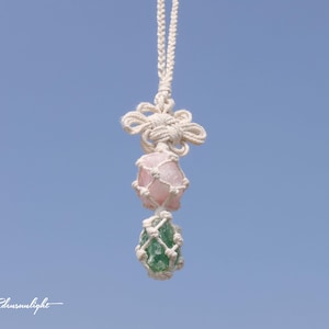 May include: A white macrame necklace with a pink and green crystal pendant. The necklace is hanging against a blue sky.