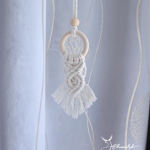 May include: A cream-coloured macrame dream catcher. It has a wooden ring, a woven centre, and a fringed base. A small wooden bead and a hanging cord are also present. The background is a light-coloured curtain.