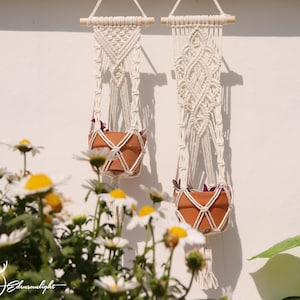 Puede incluir: Dos maceteros de macramé con macetas de terracota y plantas verdes colgando de una pared blanca. El macramé es blanco y tiene un patrón de diamante.