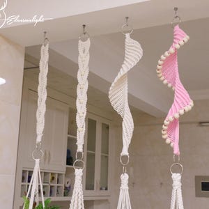 May include: Four macrame plant hangers suspended from ceiling hooks. Three hangers are made of off-white cord with varying braided and spiral knot patterns. The fourth hanger is made of pink cord with a spiral design accented by light wooden beads.