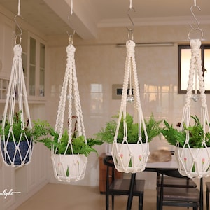 May include: Four off-white macrame plant hangers suspended by metal S-hooks, each featuring intricate knotted cord patterns and a clear spherical bead. The hangers hold potted green ferns, with two pots being white with floral designs.