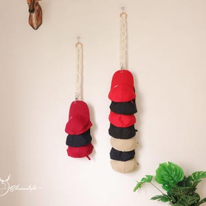 May include: Two macrame hat organizers hanging on a white wall, each holding a stack of baseball caps. The caps are in various colors, including red, black, and tan. A deer head wall decor is visible in the upper left corner.