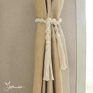 以下が含まれることがあります： クリーム色のマクラメカーテンタッセル。結び目のデザインとタッセルの端が特徴です。ベージュのカーテンに巻き付けられ、窓の装飾に装飾的なタッチを加えています。デザインには、一連の結び目と装飾的なビーズが含まれています。
