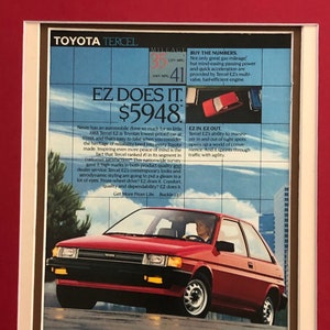 May include: A red 1988 Toyota Tercel EZ car is shown driving on a city street. The car has a white interior and a woman is driving. The ad text reads "EZ Does It. $5948. Never has an automobile done so much for so little."  The ad also highlights the car's fuel efficiency, maneuverability, and reliability.  The ad text also states that the Tercel EZ is Toyota's lowest-priced car.