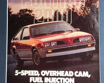 May include: A vintage print advertisement for the Pontiac 2000 car. The ad features a red and white Pontiac 2000 car with the tagline "5-Speed, Overhead Cam, Fuel Injection". The ad text highlights the car's features, including its front-wheel drive, reclining bucket seats, 5-speed manual transmission, and overhead cam 4-cylinder engine with electronic fuel injection.