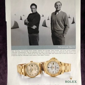 May include: Two men stand in front of a white background with shark fin shapes. The man on the left is wearing a black sweater and the man on the right is wearing a gray sweater. Both men are wearing watches. The text "Perpetual Spirit" is at the top of the image. The text "Rolex Day-Date in 18kt gold and Rolex Yacht-Master in 18kt gold" is at the bottom of the image. The text "Officially Certified Swiss Chronometers. Rolex, Oyster Perpetual, Day-Date, Yacht-Master and Perpetual Spirit are trademarks." is also at the bottom of the image. The text "FOR THE NAME AND LOCATION OF AN OFFICIAL ROLEX JEWELER NEAR YOU PLEASE CALL 1-800-36ROLEX/1-800-367-6539." is at the bottom of the image. The text "www.rolex.com" is at the bottom of the image. The text "New York" is at the bottom of the image.