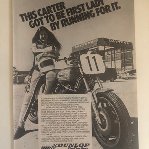 Pode incluir: Uma fotografia a preto e branco de uma mulher a correr de mota numa pista. A mota tem um pneu Dunlop. O texto "This Carter got to be first lady by running for it." est&aacute; no topo da imagem. O texto "Dunlop The Tire Pros" est&aacute; na parte inferior da imagem.