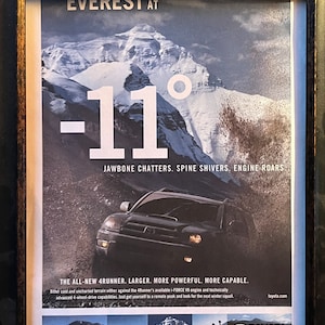 May include: Framed advertisement for the Toyota 4Runner, set against a snowy mountain backdrop. The image features the text "EVEREST AT -11°" and the tagline "JAWBONE CHATTERS. SPINE SHIVERS. ENGINE ROARS." A 4Runner is shown driving through a snowy terrain. The bottom reads "GET THE FEELING" and the Toyota logo.