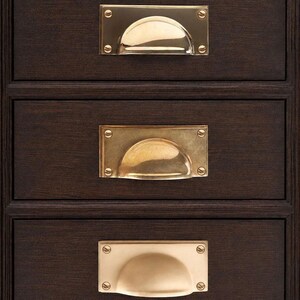 May include: A dark brown wooden cabinet featuring three horizontal drawers, each fitted with a rectangular brass cup pull handle. The handles are secured with four visible screws at the corners and exhibit varying metallic finishes.