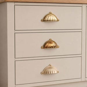 Peut inclure: Une commode en bois gris clair dotée de trois tiroirs horizontaux, chacun équipé d'une poignée coquille en laiton poli. Le meuble est surmonté d'un plateau en bois de chêne clair naturel, offrant une unité de rangement propre et fonctionnelle.
