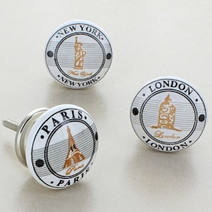 May include: Three white ceramic cabinet knobs with black and gold accents. Each knob features a different city landmark: the Statue of Liberty, Big Ben, and the Eiffel Tower. The knobs are labelled with the city names: "New York", "London", and "Paris".