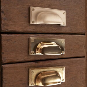 May include: A wooden cabinet featuring three horizontal drawers, each fitted with a rectangular brass cup pull handle. The handles are secured to the dark brown wood with four small screws, showcasing a polished metallic finish and curved grip.