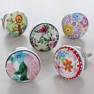 Könnte beinhalten: Fünf Keramik-Schubladengriffe mit Blumenmustern. Die Griffe sind rund und haben einen weißen Hintergrund mit bunten Blumen und Designs.