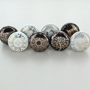 Może przedstawiać: Zestaw ośmiu ceramicznych gałek do szafek z czarnymi i złotymi wzorami kwiatowymi. Gałki są okrągłe i idealne do dodania odrobiny elegancji do każdego pomieszczenia.