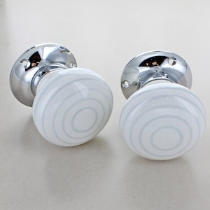 May include: Two round door knobs featuring a white ceramic surface with concentric light blue circular patterns. Each knob is attached to a polished chrome circular base plate with two visible screw holes for mounting to a door surface.