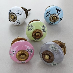 Könnte beinhalten: Fünf Keramikknöpfe mit einem abgenutzten schwarz-weißen Muster. Die Knöpfe sind in verschiedenen Farben: weiß, hellblau, hellgrün, rosa und grau. Jeder Knopf hat eine Schraube aus Messing und einen Messingring um die Mitte.