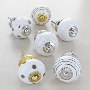 Op de afbeelding: Zes witte keramische kastknoppen met gouden en zilveren accenten. De knoppen hebben verschillende patronen, waaronder stippen, strepen en een gebarsten afwerking.