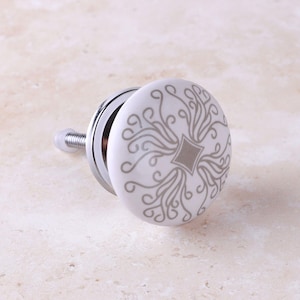 May include: A round white ceramic cabinet knob featuring a central diamond shape surrounded by intricate gray swirling line patterns. The knob is attached to a silver-toned metal base with a threaded bolt extending from the back for installation.