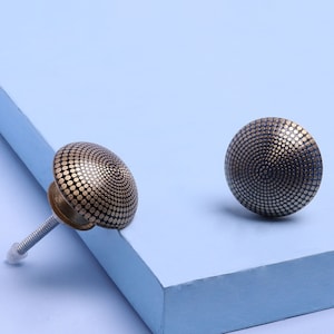 Puede incluir: Dos pomos de armario dorados con un diseño estampado en blanco y negro. Los pomos son redondos y tienen un centro elevado.