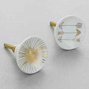 May include: Two white ceramic cabinet knobs with gold accents. One knob features a sunburst design, while the other has three gold arrows.