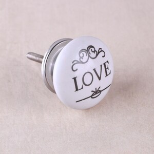 Può includere: Un pomello rotondo per mobili in ceramica bianca con un design stampato in nero che riporta la parola LOVE in lettere maiuscole, decorato con volute sopra e una linea orizzontale con un piccolo fiocco sotto, fissato a un bullone in metallo.