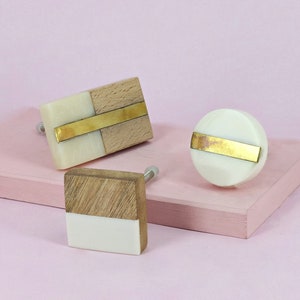 May include: Three decorative cabinet knobs with a white and wood design. Each knob features a gold metal strip across the centre. The knobs are on a pink surface.