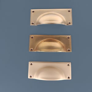 Può includere: Tre maniglie per cassetti rettangolari in ottone disposte verticalmente. Ogni maniglia presenta una conca centrale incassata e quattro fori circolari per le viti, uno per ogni angolo. Le superfici metalliche mostrano varie tonalità dorate.