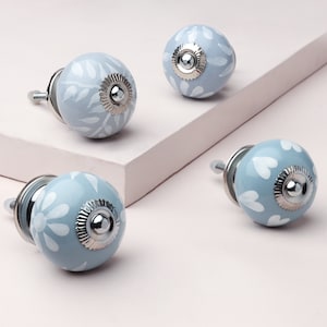 Könnte beinhalten: Vier Keramik-Schrankknöpfe mit hellblauem Hintergrund und weißen Blumenmustern. Die Knöpfe haben eine silberne Metallbasis und eine Schraube.