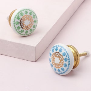 Peut inclure: Deux boutons de cabinet en céramique avec des motifs floraux dorés et bleus ou verts. Les boutons ont une vis dorée et sont fixés à une surface blanche.