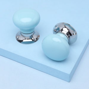 May include: Two light blue ceramic door knobs with a glossy finish. The knobs have a rounded shape and are mounted on a shiny, silver-colored metal base with screw holes. The background is a light blue surface.
