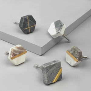 Puede incluir: Cinco pomos de armario de mármol diferentes con detalles dorados. Los pomos son de diferentes formas y tamaños, con una mezcla de mármol gris, blanco y marrón.