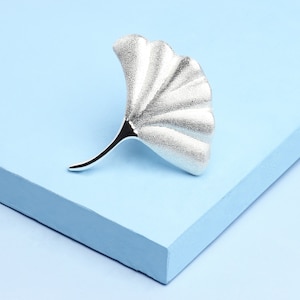 May include: A silver ginkgo leaf brooch with a textured surface. The brooch is on a light blue background.