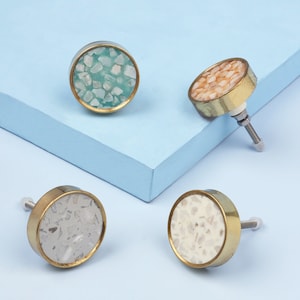 May include: Four round cabinet knobs with a gold metal base and a decorative top made of different colored stone chips. The knobs are on a light blue surface.