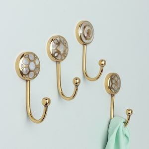 May include: Four gold-toned hooks with decorative white and gold inlays. The hooks are mounted on a light green wall.