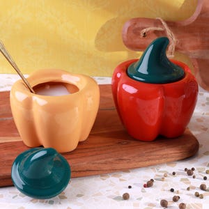 Op de afbeelding: Twee keramische pepervormige containers. Eén is oranje met een lepel erin, de andere is rood met een groen deksel. Een los groen deksel ligt in de buurt. De containers staan op een houten snijplank.