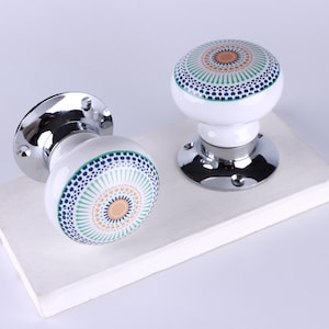 May include: Two white ceramic door knobs with a decorative circular pattern in blue, green, and tan. The knobs have a chrome base and are displayed on a white surface. The knobs are a decorative home decor item.