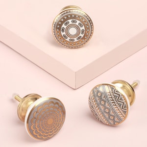 May include: Three decorative cabinet knobs with brass hardware. The knobs feature intricate patterns in gold, white, and gray. One knob has a floral design, while the others have geometric patterns. The knobs are set against a light pink background.