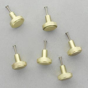May include: Six brass cabinet knobs with a textured, circular design. Each knob has a silver-colored screw post. The knobs are arranged on a light gray surface.