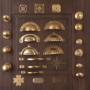 May include: A collection of brass hardware mounted on a dark wood panel, featuring various drawer pulls, knobs, and decorative plates. The assortment includes semi-circular cup pulls, straight bar handles, round knobs, and geometric shapes.