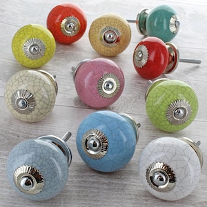May include: A selection of ceramic cabinet knobs in various colours. Each knob has a crackle glaze finish and a silver-toned metal base. The colours include red, yellow, green, pink, blue, and white. These knobs are likely for furniture or decorative purposes.