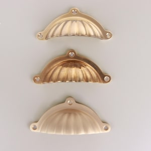 May include: Three semi-circular metal cup pulls arranged vertically. Each handle features a scalloped, ribbed design with three mounting holes, one at the top center and two at the bottom corners. The finishes vary in tone from bright gold to brass.