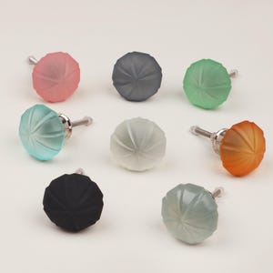 Könnte beinhalten: Acht farbige Glasschrankknöpfe in verschiedenen Farben. Die Knöpfe sind in Rosa, Grau, Grün, Blau, Weiß, Orange, Schwarz und Graugrün gehalten. Jeder Knopf hat ein radiales Design und eine silberne Schraube.