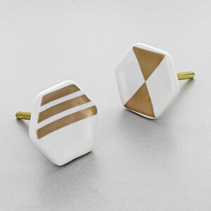 May include: Two white and gold geometric cabinet knobs. One knob features three horizontal gold stripes, while the other has a gold triangle design. Both have brass screws for installation. These decorative knobs add a modern touch to furniture.