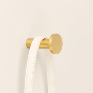 May include: A gold-colored metal hook with a cylindrical body and a circular head. A white leather strap is hanging from the hook. The hook is mounted on a white wall.
