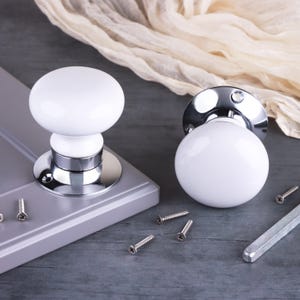 May include: Two round white ceramic cabinet knobs with polished chrome bases. One knob is mounted on a grey panel, while the other sits beside it. The set includes several small metal screws and a rectangular metal spindle for installation.