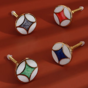 May include: Four round cabinet knobs with a gold finish. Each knob has a white centre with a different coloured geometric design: blue, green, red, and grey.