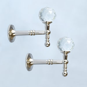 Könnte beinhalten: Zwei dekorative Wandhaken mit silbernem Finish und klaren Kristallkugel-Abschlüssen. Die Haken haben einen runden Sockel und einen dekorativen Stiel. Die Kristallkugeln sind facettiert und reflektieren das Licht.